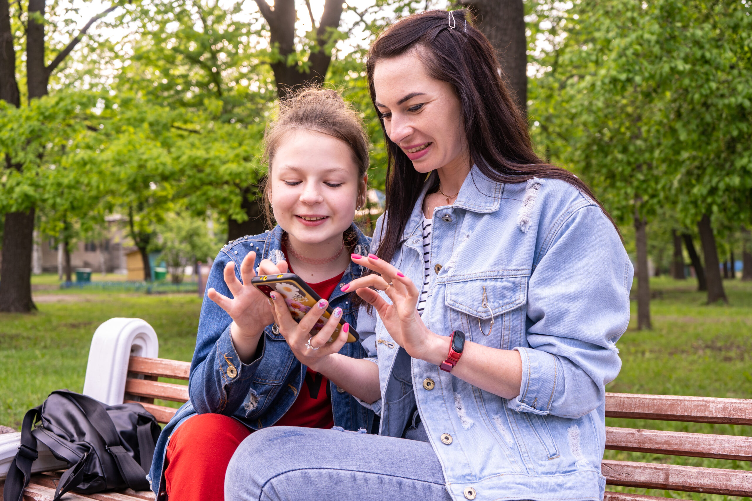 Mother and teenage daughter walking in a park, sitting on a bench and looking in smartphone, happy young caucasian woman with long hair and teenage girl surfing the internet outdoors, lifestyle family